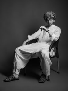 Gillian as a man in a white salwar kameez sitting arrogantly in a chair